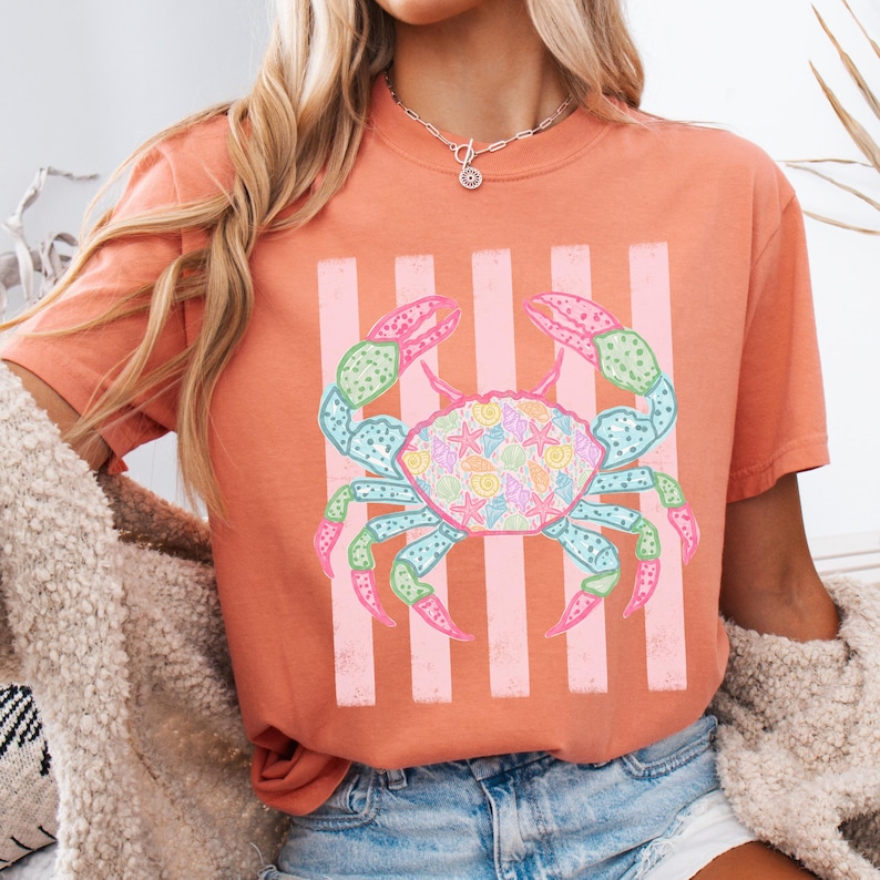 May include: Coral-colored t-shirt featuring a colorful crab design. The crab has blue, green, pink, and yellow accents, with a shell filled with seashell patterns. Vertical pink stripes are behind the crab.