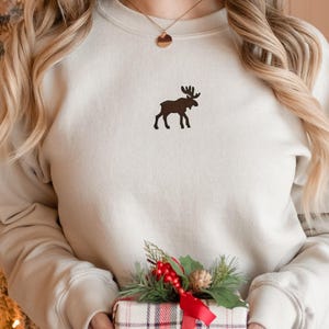 May include: A cream-colored sweatshirt with a brown moose silhouette embroidered on the front. The sweatshirt is being held by a person wearing a gold necklace.