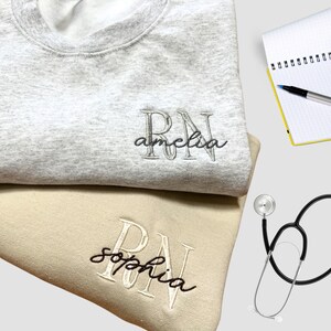 Puede incluir: Dos sudaderas grises y beige con las letras "RN" bordadas. Las sudaderas tienen los nombres "Amelia" y "Sophia" bordados debajo de las letras.