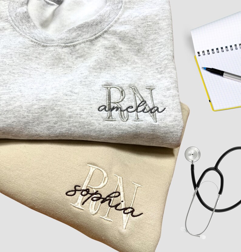 Puede incluir: Dos sudaderas grises y beige con las letras "RN" bordadas. Las sudaderas tienen los nombres "Amelia" y "Sophia" bordados debajo de las letras.