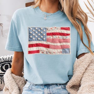 K&ouml;nnte beinhalten: Hellblaues T-Shirt mit einem Patchwork-Design der amerikanischen Flagge. Die Flagge zeigt wei&szlig;e Sterne auf blauem Grund und rote und gemusterte Streifen. Das T-Shirt ist aus weichem Material und hat kurze &Auml;rmel.