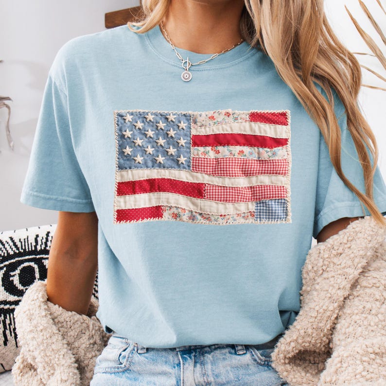 K&ouml;nnte beinhalten: Hellblaues T-Shirt mit einem Patchwork-Design der amerikanischen Flagge. Die Flagge zeigt wei&szlig;e Sterne auf blauem Grund und rote und gemusterte Streifen. Das T-Shirt ist aus weichem Material und hat kurze &Auml;rmel.