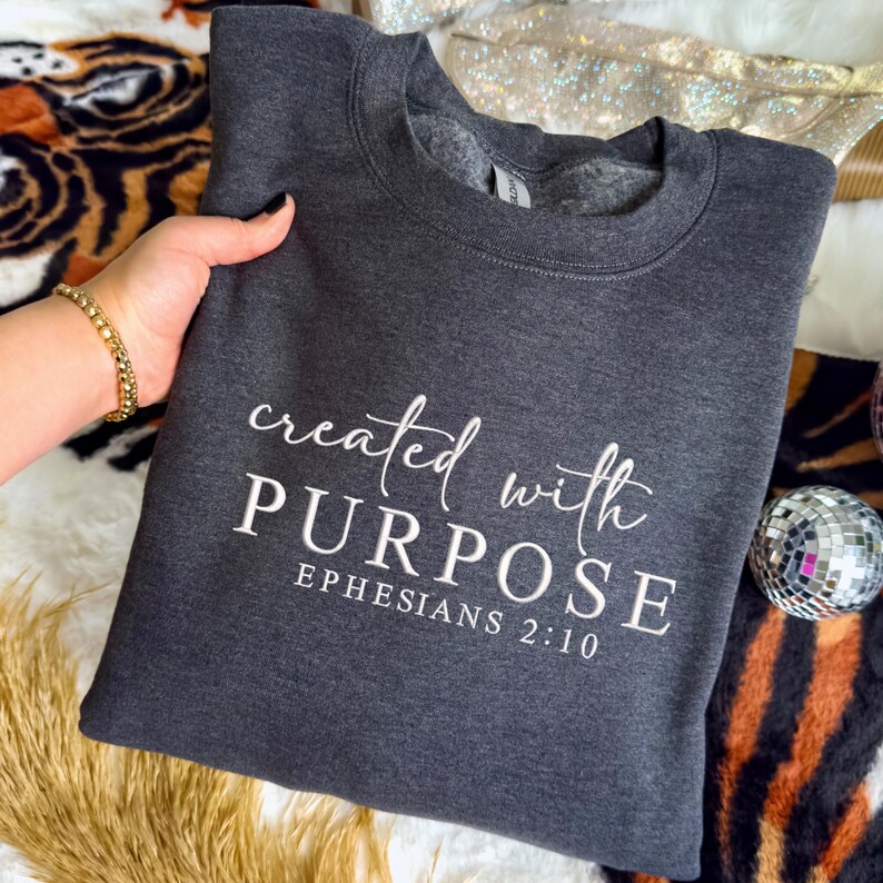May include: A dark grey sweatshirt featuring the white text "created with PURPOSE" and "EPHESIANS 2:10". The garment is displayed on a patterned surface.