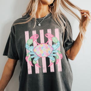 May include: Dark gray t-shirt featuring a colorful crab design. The crab is depicted with pink, blue, green, and yellow accents, set against a backdrop of pink vertical stripes. The crab's shell is filled with seashell illustrations.
