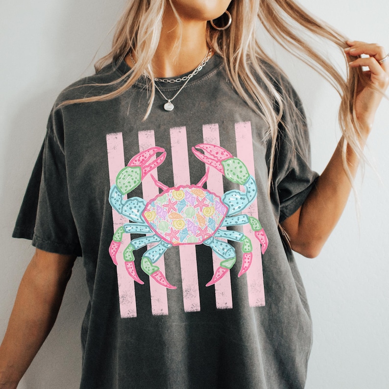 May include: Dark gray t-shirt featuring a colorful crab design. The crab is depicted with pink, blue, green, and yellow accents, set against a backdrop of pink vertical stripes. The crab's shell is filled with seashell illustrations.