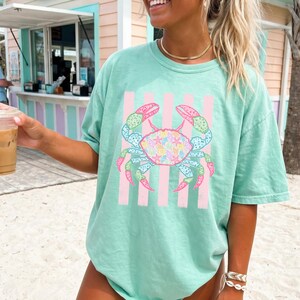 May include: A light green t-shirt featuring a colorful crab design with pink vertical stripes. The crab has a variety of colors, including blue, pink, green, and yellow. The crab's body is filled with seashell patterns. The shirt is a relaxed fit.
