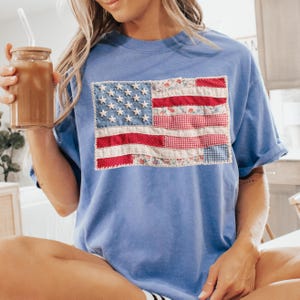 K&ouml;nnte beinhalten: Hellblaues T-Shirt mit einem Patchwork-Design der amerikanischen Flagge. Die Flagge verwendet rote, wei&szlig;e und blaue Stoffe, einschlie&szlig;lich Blumen- und Vichy-Mustern. Das T-Shirt ist ein l&auml;ssiger Stil.