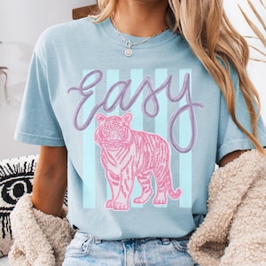K&ouml;nnte beinhalten: Hellblaues T-Shirt mit einem rosa Tiger-Motiv und dem Wort "Easy" in Schreibschrift. Der Tiger steht vor hellblauen vertikalen Streifen. Das T-Shirt ist aus weichem Material.