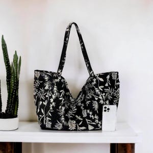 May include: A black tote bag with a white floral pattern. The bag has two long handles and is made of a fabric material. A white phone is sitting inside the bag.