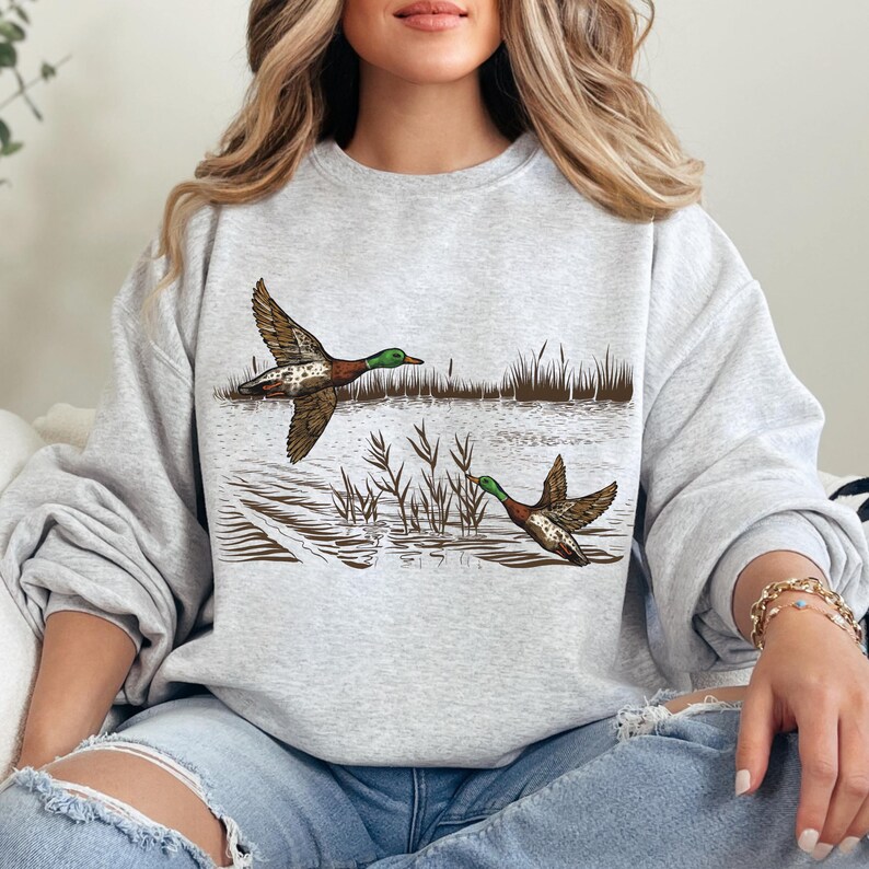K&ouml;nnte beinhalten: Ein hellgraues Sweatshirt mit einer detaillierten Illustration von zwei Enten im Flug &uuml;ber Wasser und Schilf. Die Enten haben braune, gr&uuml;ne und wei&szlig;e Markierungen. Das Design ist eine Grafik zum Thema Natur.