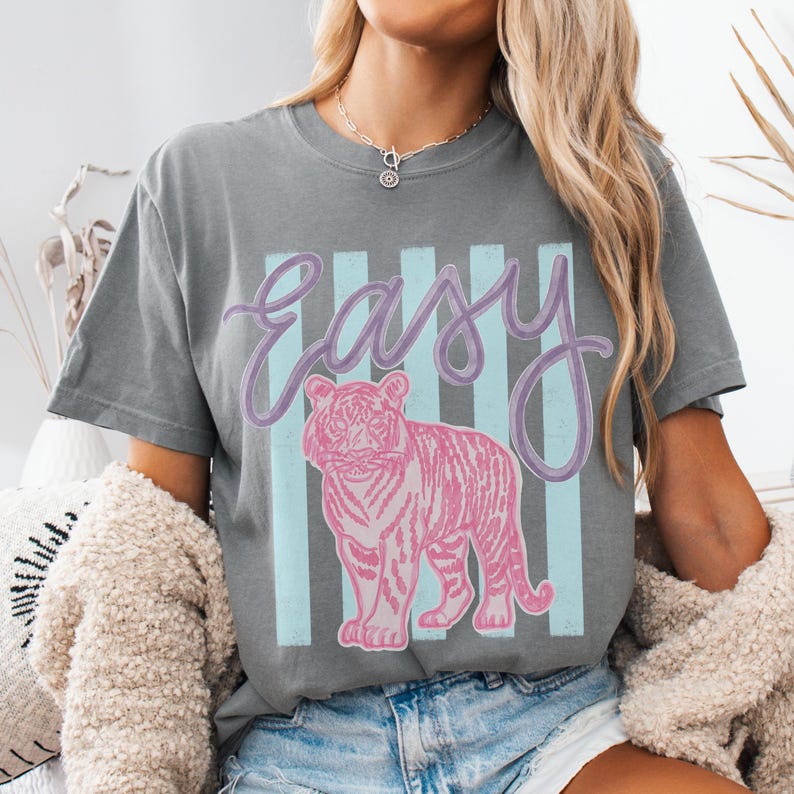 K&ouml;nnte beinhalten: Graues T-Shirt mit dem Wort "Easy" in Schreibschrift und einer rosa Tigergrafik. Das Wort "Easy" ist lavendelfarben, und der Tiger ist rosa mit Streifen. Der Hintergrund hat hellblaue vertikale Streifen.