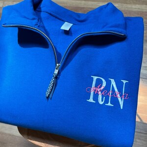 May include: A royal blue quarter-zip sweatshirt with white embroidered "RN" and the name "Alexa" in pink script. The sweatshirt has a silver zipper and a folded collar. The garment is made of a soft material and is folded on a wooden surface.