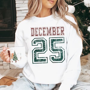 Pu&ograve; includere: Felpa bianca con la scritta "DECEMBER 25" in un carattere invecchiato in stile collegiale. La parola "DECEMBER" &egrave; marrone e il numero "25" &egrave; verde. Un capo d'abbigliamento festivo.