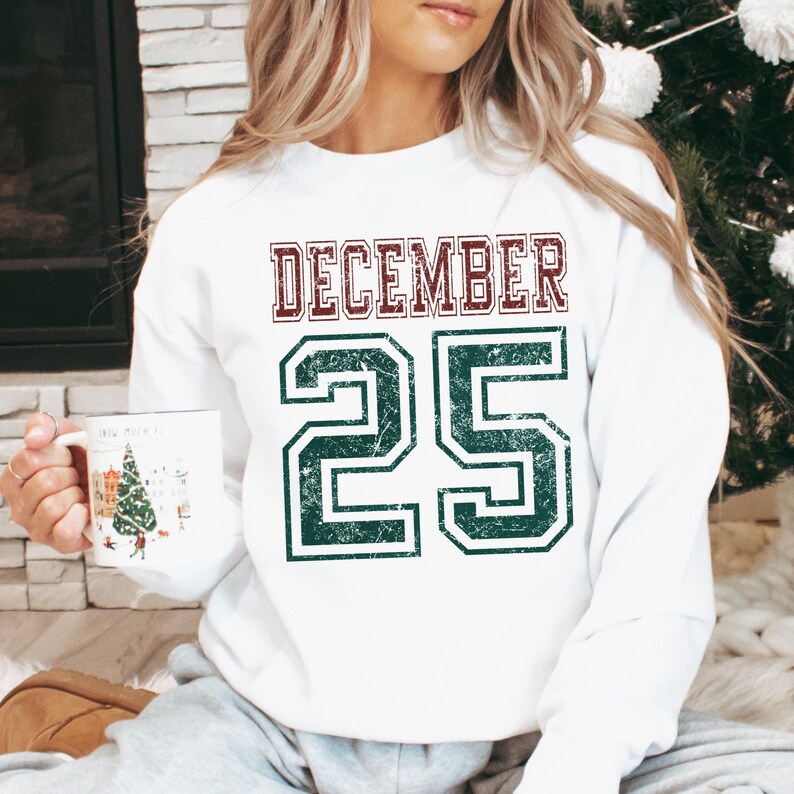 Pu&ograve; includere: Felpa bianca con la scritta "DECEMBER 25" in un carattere invecchiato in stile collegiale. La parola "DECEMBER" &egrave; marrone e il numero "25" &egrave; verde. Un capo d'abbigliamento festivo.