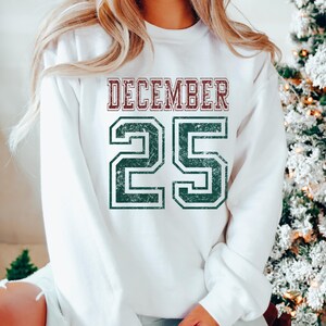 Pu&ograve; includere: Felpa bianca con la scritta "DECEMBER 25" in un carattere invecchiato in stile collegiale. La parola "DECEMBER" &egrave; color amaranto e il numero "25" &egrave; verde. Un capo d'abbigliamento festivo.