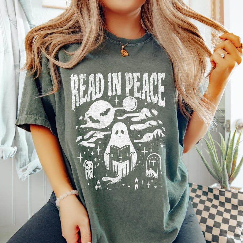 May include: Dark green t-shirt with the text "READ IN PEACE" in a distressed white font. The graphic features a ghost reading a book, bats, a moon, and a graveyard scene. The shirt is a comfortable, casual style.