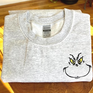 Puede incluir: Una sudadera gris claro con cuello redondo. La sudadera presenta una cara de Grinch bordada con ojos amarillos y una sonrisa negra. La etiqueta de la marca Gildan es visible. La sudadera est&aacute; doblada sobre una superficie de madera.