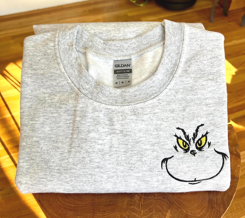 Puede incluir: Una sudadera gris claro con cuello redondo. La sudadera presenta una cara de Grinch bordada con ojos amarillos y una sonrisa negra. La etiqueta de la marca Gildan es visible. La sudadera est&aacute; doblada sobre una superficie de madera.