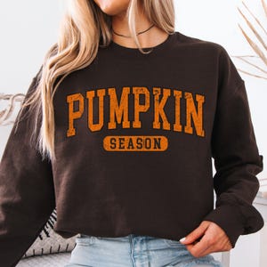 May include: A brown sweatshirt with the words "PUMPKIN SEASON" in orange block letters. The text is slightly distressed, giving it a vintage look. The sweatshirt has a crew neck and long sleeves.