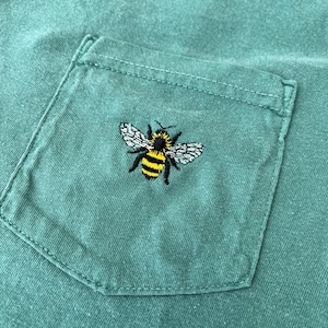 May include: A sage green t-shirt featuring a chest pocket with a small, detailed embroidery of a bumblebee. The bee is depicted with black and yellow horizontal stripes on its body, black legs, and textured white wings centered on the pocket.