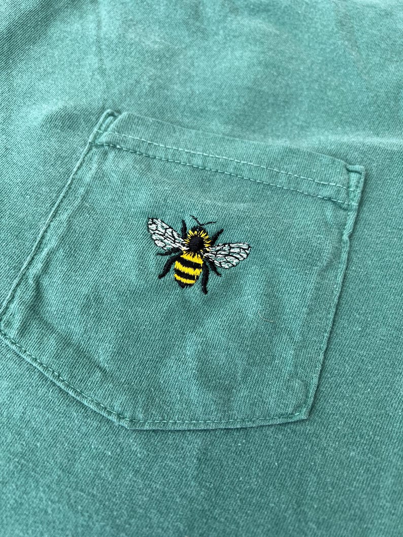 May include: A sage green t-shirt featuring a chest pocket with a small, detailed embroidery of a bumblebee. The bee is depicted with black and yellow horizontal stripes on its body, black legs, and textured white wings centered on the pocket.