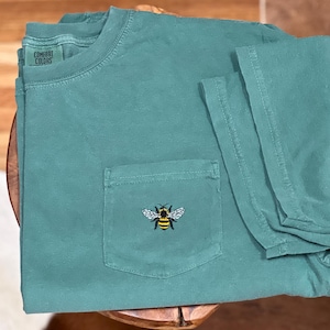 May include: A teal green short-sleeve t-shirt with a crew neckline and a single chest pocket. The shirt features an embroidered bee graphic on the pocket and a Comfort Colors brand label printed on the inside of the collar area.