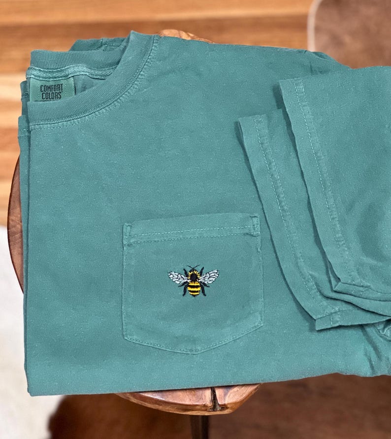 May include: A teal green short-sleeve t-shirt with a crew neckline and a single chest pocket. The shirt features an embroidered bee graphic on the pocket and a Comfort Colors brand label printed on the inside of the collar area.