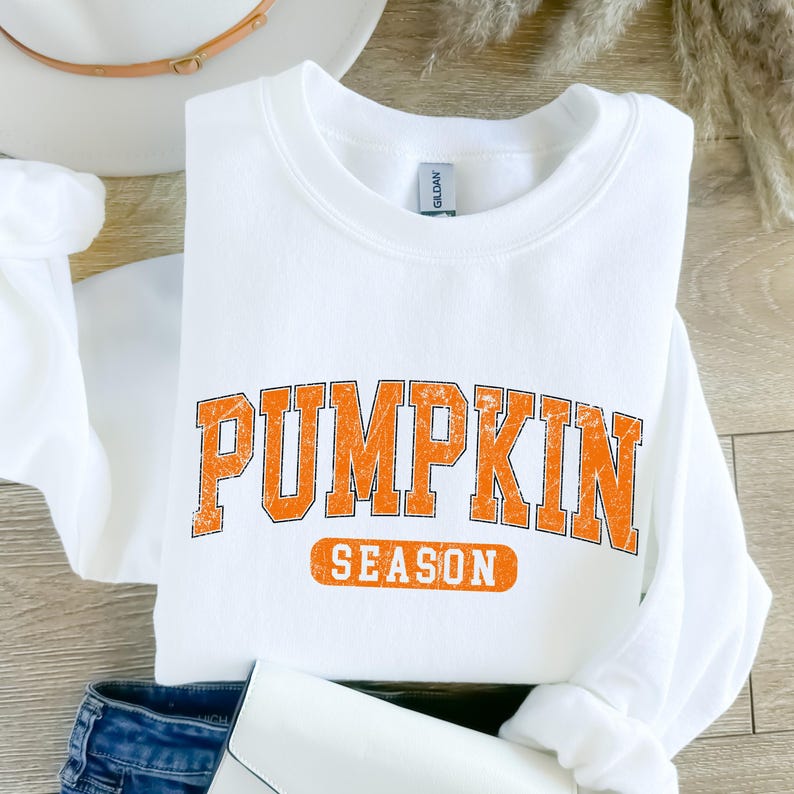May include: A white sweatshirt with the words "PUMPKIN SEASON" printed in orange block letters. The sweatshirt is folded, with a pair of blue jeans and a white clutch visible in the background. A white hat is also in the background.