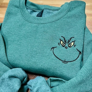 Puede incluir: Sudadera verde con un rostro del Grinch bordado, con ojos amarillos y una sonrisa negra. La sudadera de cuello redondo tiene mangas largas. La tela es un material suave y jaspeado.