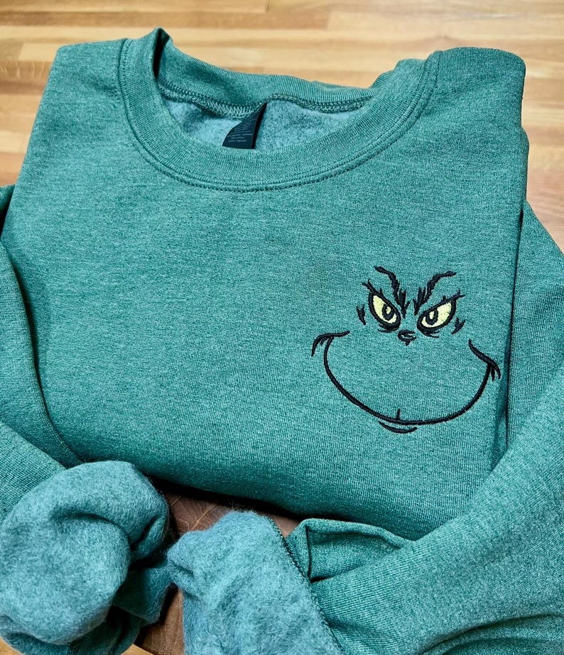 Puede incluir: Sudadera verde con un rostro del Grinch bordado, con ojos amarillos y una sonrisa negra. La sudadera de cuello redondo tiene mangas largas. La tela es un material suave y jaspeado.