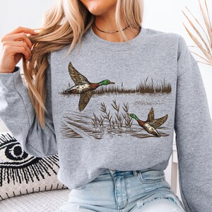 K&ouml;nnte beinhalten: Ein graues Sweatshirt mit einer detaillierten Illustration von zwei Enten im Flug &uuml;ber Wasser. Die Enten haben braune, wei&szlig;e und gr&uuml;ne Markierungen mit ausgebreiteten Fl&uuml;geln. Schilf und Wasserelemente vervollst&auml;ndigen das Naturthema.
