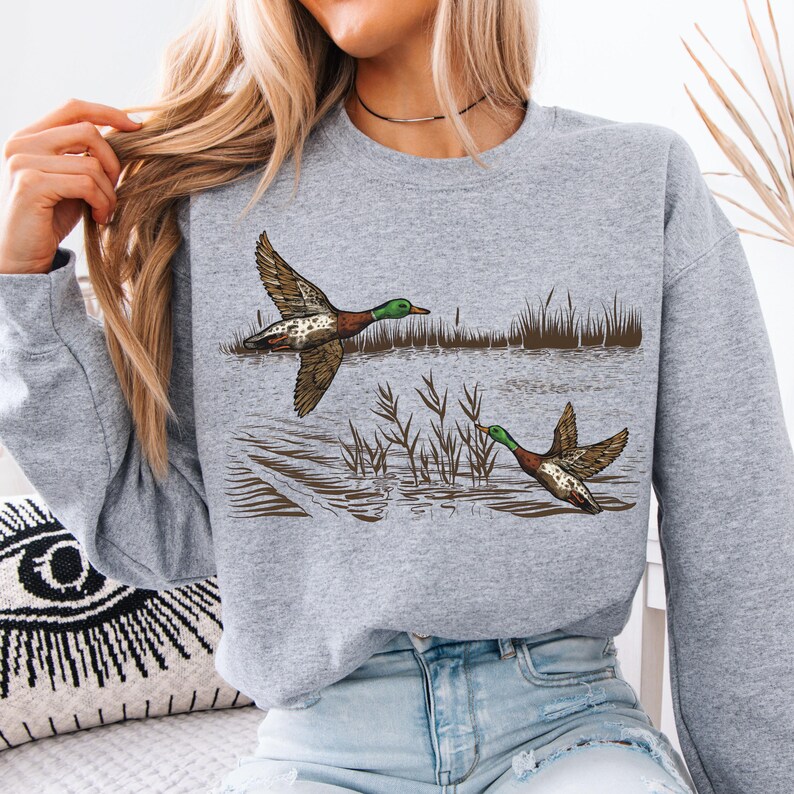 K&ouml;nnte beinhalten: Ein graues Sweatshirt mit einer detaillierten Illustration von zwei Enten im Flug &uuml;ber Wasser. Die Enten haben braune, wei&szlig;e und gr&uuml;ne Markierungen mit ausgebreiteten Fl&uuml;geln. Schilf und Wasserelemente vervollst&auml;ndigen das Naturthema.