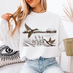 K&ouml;nnte beinhalten: Wei&szlig;es Sweatshirt mit einer detaillierten Illustration von zwei Enten im Flug &uuml;ber Wasser und Schilf. Die Enten sind braun, gr&uuml;n und wei&szlig;. Das Sweatshirt ist lang&auml;rmlig und hat einen Rundhalsausschnitt.