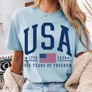 May include: Light blue t-shirt with the word "USA" in navy blue block letters. Below the word "USA" is the American flag, the years 1776 and 2026, and the text "250 YEARS OF FREEDOM."