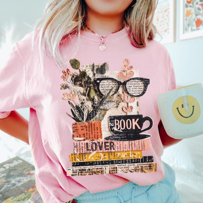 May include: Pink t-shirt with a collage design featuring glasses, a coffee cup, and the word "BOOK" and "LOVER". The design includes floral elements and text fragments. A mug with a smiley face is held in the image.