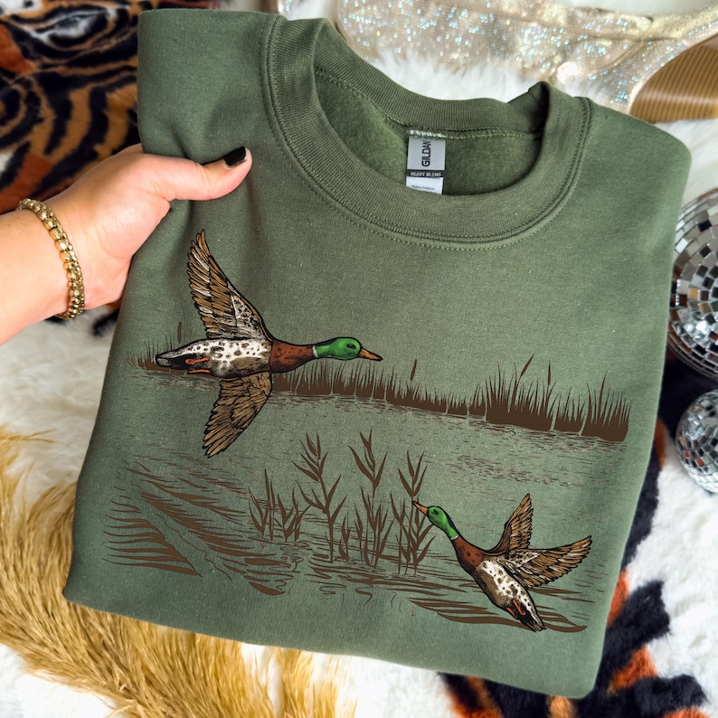 K&ouml;nnte beinhalten: Ein olivgr&uuml;nes Sweatshirt mit einer detaillierten Illustration von zwei Enten im Flug &uuml;ber Wasser und Schilf. Die Enten haben braune, wei&szlig;e und gr&uuml;ne Markierungen. Ein bequemes, l&auml;ssiges Kleidungsst&uuml;ck.