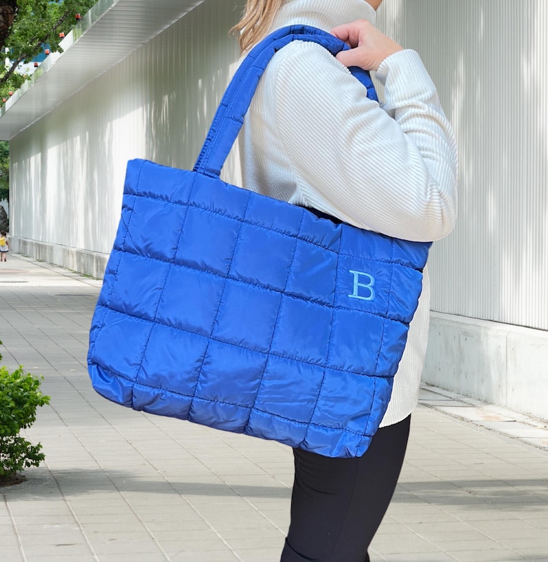 May include: A vibrant blue quilted tote bag with a stitched grid pattern. The bag features a matching blue handle and a large letter "B" embroidered on the front. The bag is made of a soft, padded material.
