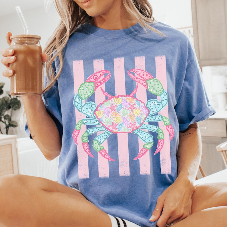 May include: A blue t-shirt featuring a colorful crab design. The crab is decorated with seashells and starfish, set against pink vertical stripes. The shirt has short sleeves and a relaxed fit, perfect for a casual summer look.
