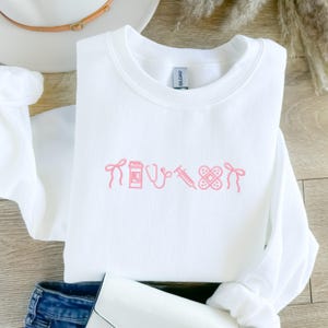 Pode incluir: Camisola branca com s&iacute;mbolos m&eacute;dicos rosa bordados, incluindo um la&ccedil;o, um frasco de comprimidos, um estetosc&oacute;pio, uma seringa e um penso. A camisola est&aacute; dobrada, com um chap&eacute;u e cal&ccedil;as de ganga no fundo.