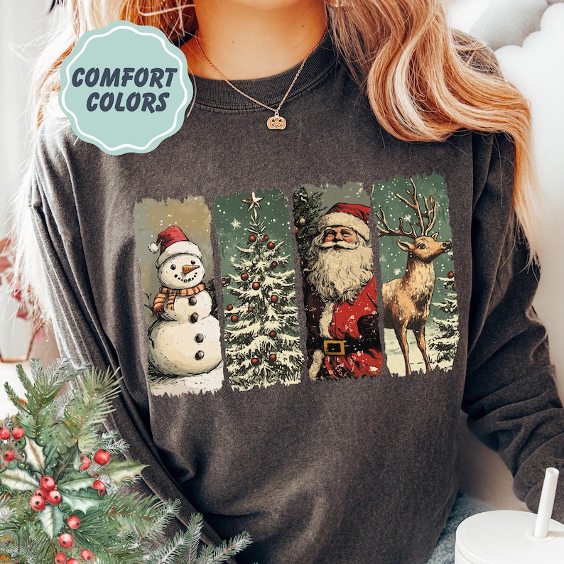 May include: Dark grey sweatshirt with a Christmas-themed graphic. The graphic features a snowman, Christmas tree, Santa Claus, and a reindeer. The text "COMFORT COLORS" is in a light blue circle in the upper left corner.