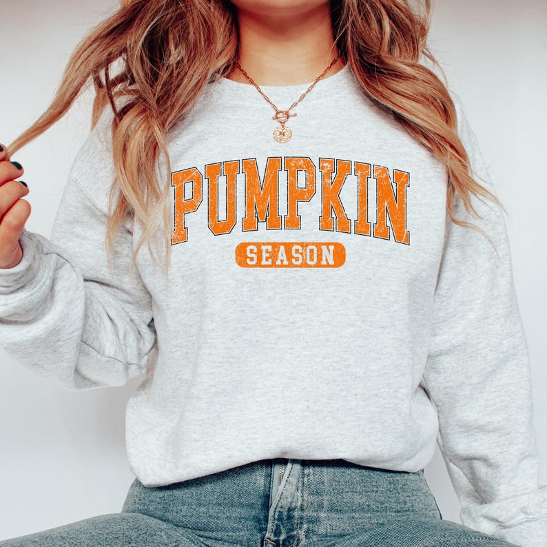 May include: A light gray sweatshirt with the words "PUMPKIN SEASON" in orange block letters. The sweatshirt has a crew neck and long sleeves. The text is in a collegiate style font, perfect for fall.
