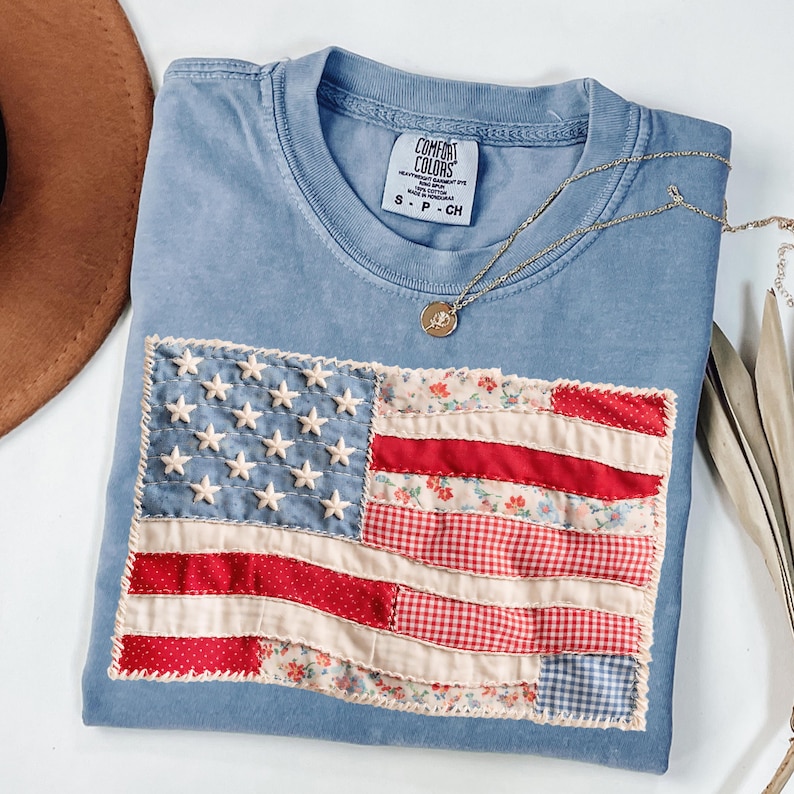 K&ouml;nnte beinhalten: Ein hellblaues Comfort Colors T-Shirt mit einem aufgen&auml;hten amerikanischen Flaggen-Design. Die Flagge hat wei&szlig;e Sterne, rot-wei&szlig;e Streifen und Blumen- und Vichy-Muster. Eine goldene Halskette mit Anh&auml;nger liegt auf dem T-Shirt.