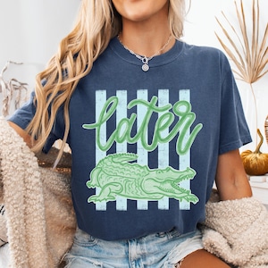 Puede incluir: Camiseta azul marino con un gr&aacute;fico de cocodrilo verde y la palabra "later" en cursiva. La palabra "later" est&aacute; encima del cocodrilo, con rayas verticales de color azul claro detr&aacute;s. La camiseta es de estilo casual.