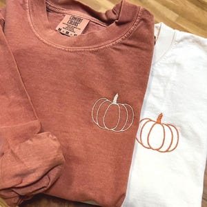 May include: A terracotta long-sleeve shirt with a white embroidered pumpkin outline and a white long-sleeve shirt with an orange embroidered pumpkin outline. Both shirts feature a Comfort Colors label at the inner neckline with size M.