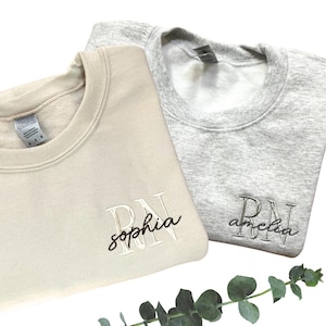 Puede incluir: Dos sudaderas de color crema y gris con las letras RN bordadas. La sudadera de color crema tiene el nombre "sophia" bordado debajo de las letras. La sudadera gris tiene el nombre "amelia" bordado debajo de las letras.