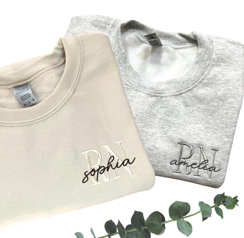 Puede incluir: Dos sudaderas de color crema y gris con las letras RN bordadas. La sudadera de color crema tiene el nombre "sophia" bordado debajo de las letras. La sudadera gris tiene el nombre "amelia" bordado debajo de las letras.