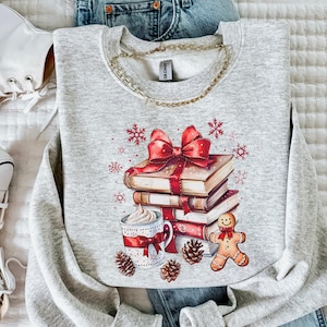 May include: A light grey sweatshirt with a graphic print. The graphic includes a stack of books tied with a red bow, a mug of whipped cream, a gingerbread man, pine cones, and snowflakes. A gold chain is around the neckline.