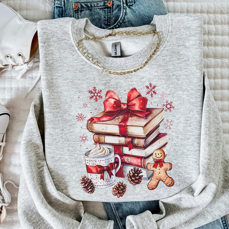 May include: A light grey sweatshirt with a graphic print. The graphic includes a stack of books tied with a red bow, a mug of whipped cream, a gingerbread man, pine cones, and snowflakes. A gold chain is around the neckline.