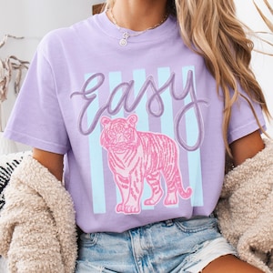 K&ouml;nnte beinhalten: Ein lila T-Shirt mit einer rosa Tiger-Illustration und dem Wort "Easy" in Schreibschrift. Das Design umfasst hellblaue vertikale Streifen, die eine l&auml;ssige und verspielte &Auml;sthetik schaffen. Das T-Shirt hat eine lockere Passform.