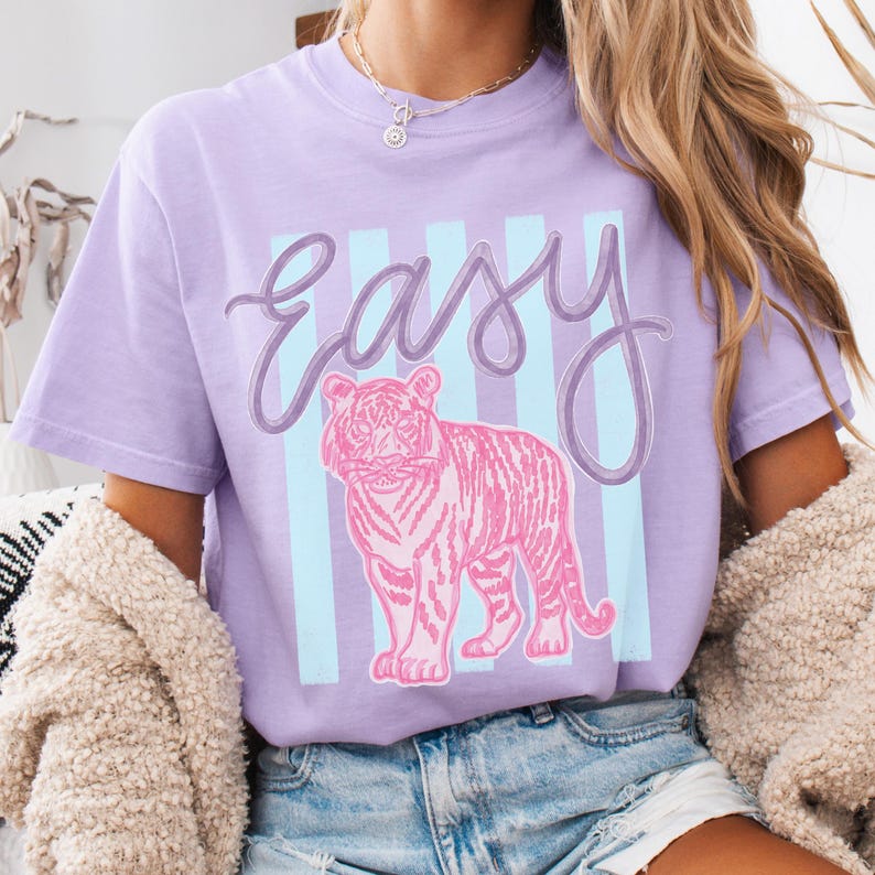 K&ouml;nnte beinhalten: Ein lila T-Shirt mit einer rosa Tiger-Illustration und dem Wort "Easy" in Schreibschrift. Das Design umfasst hellblaue vertikale Streifen, die eine l&auml;ssige und verspielte &Auml;sthetik schaffen. Das T-Shirt hat eine lockere Passform.
